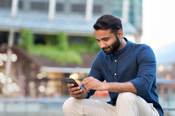 Smiling indian businessman, eastern business man entrepreneur holding cell phone using smartphone mobile tech app texting, reading news in chats on cellphone sitting on urban city street outdoor.