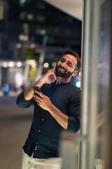 Happy smiling indian eastern man wearing earbuds holding cell phone using online audio tracks streaming smartphone application enjoying listening to new mobile dj music mix at night outdoor, vertical.