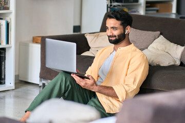Happy indian man wearing earbud sitting at home relaxing using laptop looking at pc computer...