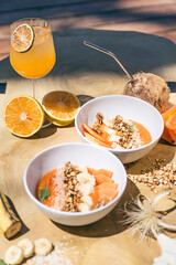 Vegan bowl. Papaya, banana, granola and coconut in a white bowl. Orange juice in a glass tumbler. All on a wooden table. Summer vibes