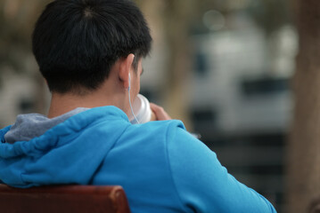 Asian man with blue hoodie listens to music with earphones as he drinks a cup of coffee outside a cafe in the middle of the day. Photo from the back.