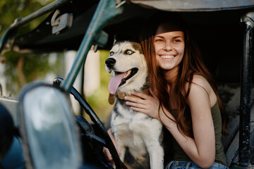 A beautiful young woman sits behind the wheel of her car together with a husky dog ​​and smiles cheerfully rejoices at a trip to nature in the evening at sunset