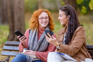 Cheerful female friends sitting on a bench in a park talking, smiling, and using smartphones
