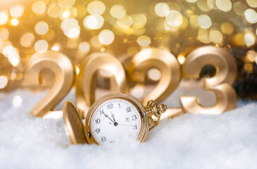 The golden watch lies on the snow against the background of the golden numbers 2023 with a golden...