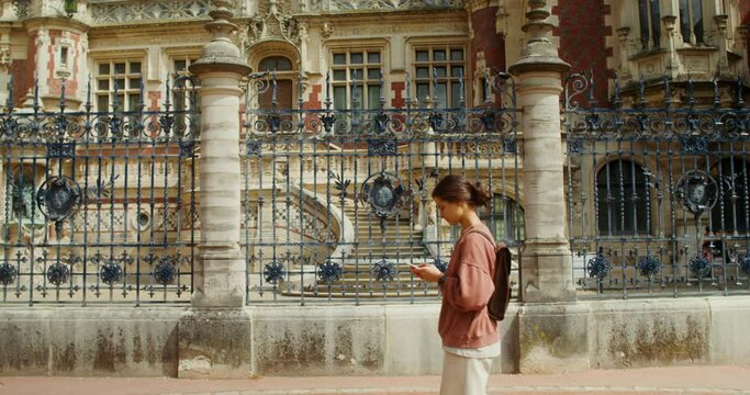 A beautiful young woman uses a smartphone while walking through a beautiful European city passing by the Benedictine Palace, F camp, France. 4k video, red komodo
