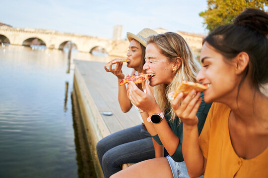 Fototapeta Three happy women sitting on the lakeside of the city eating pizza in a street stall. The happy girls enjoy the weekend together outdoors.