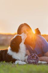 Woman and Border Collie dog licking her face lying on the grass in the park during golden hour