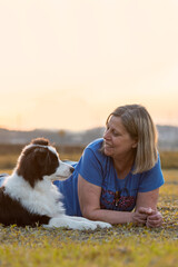 Woman and Border Collie dog looking at each other lying on the grass in the park during golden hour
