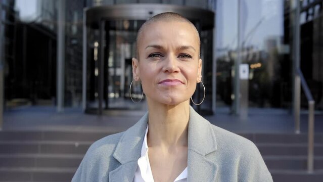 Cheerful middle-aged empowered woman with shaved hair looking at camera. Business people outdoors on background with buildings. corporate.
