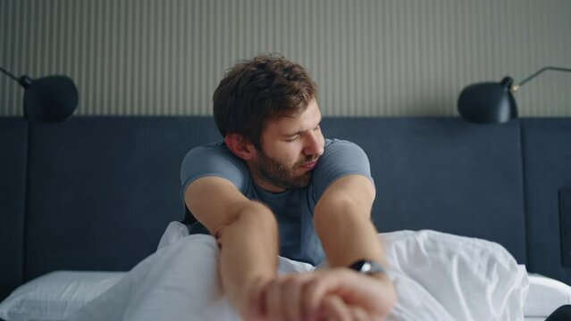 handsome man is waking up alone in bed in morning, sitting on bed, yawning and looking at smartwatch
