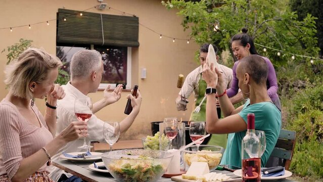 Middle aged people having dinner and fun drinking red wine together at home garden. Happy friends eating bbq food in backyard. Lifestyle of friendship and community