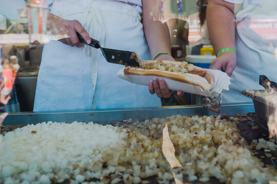 Worker scoops grilled onions onto a footlong hotdog while working in a booth at a fair