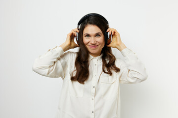 an emotional, beautiful, pleasant woman stands in a white shirt listening to music and holding headphones with her hands, smiling pleasantly with pleasure.