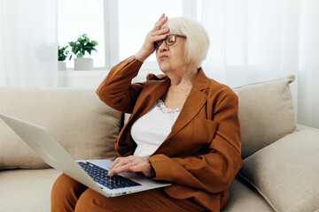 a sad elderly woman with problems at work is sitting at home on the sofa opposite the window and holding her hand near her face in a frustrated gesture looks at the laptop monitor