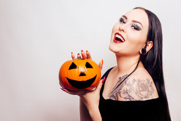 A beautiful brunette holds a pumpkin in her hands and looks at it. Woman with Halloween makeup, dark lipstick. Model in white