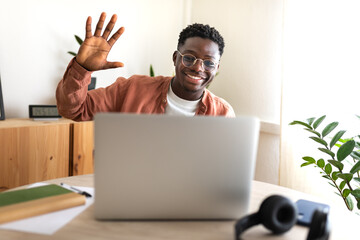 Black young man in video call taking online classes. Greeting classmates. Waving hand towards laptop. Working from home