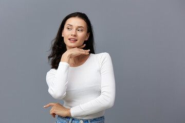Obraz premium a sophisticated woman stands on a gray background in a white T-shirt, looks mysteriously to the side, touching her chin with her hand, putting her other hand in front of her