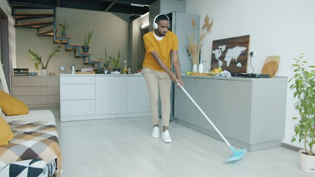 Guy wearing headphones mopping floor in kitchen and dancing with plastic mop having fun doing housework activities. Lifestyle and home concept.