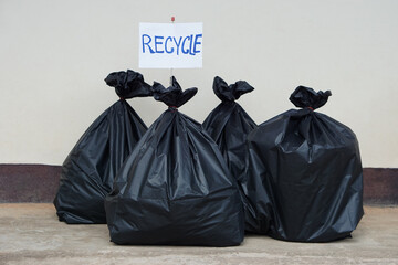 Pile of black plastic bags that contain garbage inside. Paper sign with word " RECYCLE". Concept : Waste management and sorting. Environment issue. Some rubbish can be recycled. 