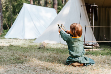 Little boy in medieval rustic clothes playing in forest. Child shooting from slingshot sitting on...