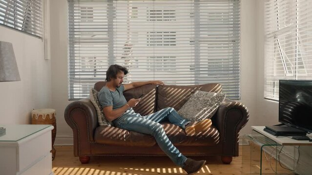 Man scrolling on phone lying on sofa. Comfortable home interior.