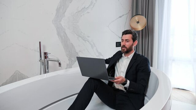 Funny young businessman lies in bath relaxes in luxurious bathroom having video call using laptop app. Satisfied attractive man answers friendly call chatting with friend uses computer indoor.