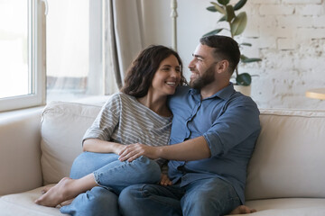 Beautiful millennial couple in love sit on sofa enjoy weekend at modern home, talking, feel happy....