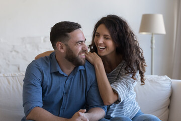 Joyful laughing couple in love sit on sofa in living room. Hispanic wife hugs husband enjoy carefree cheery conversation having pleasant talk spend weekend at home. Relations, happy marriage concept