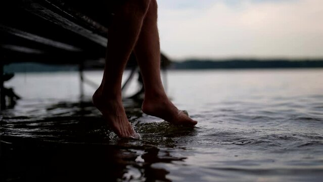 person is sitting on wooden pier adn swaying bare feet in water of sea or river in dawn or sunset