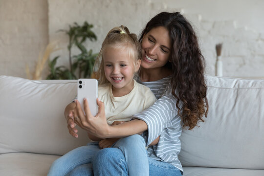 Cute girl her lovely young mother make selfie pictures on cell phone sit on sofa have fun at home using new mobile app cool funny filters, record video for new vlog looking happy. Modern tech concept