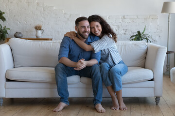 Multi ethnic attractive couple in love hugging seated on sofa at home laughing enjoy dating, spend carefree weekend together. Homeowners family portrait, relationships, happy marriage, tenancy concept