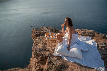 Woman covered with a blanket of bed relaxing and watching the seascape at sunrise. She holds a cup of coffee in her hand in front of her is a table with fruits and croissants. Wanderlust and freedom