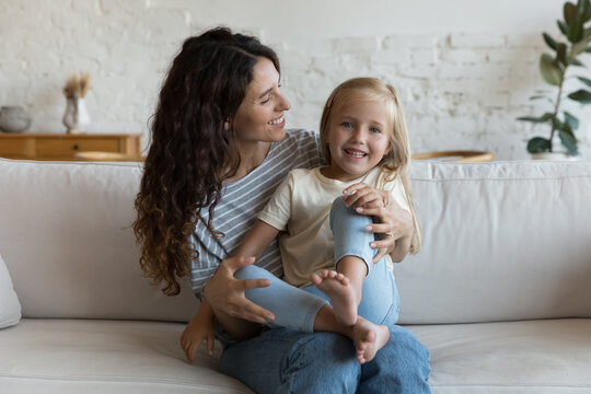 Little 5s cute blond daughter sits on loving Hispanic mothers laps smile looking at camera, enjoy time, tender moments together seated on sofa at home. Happy family, adoption, bonding, cherish concept