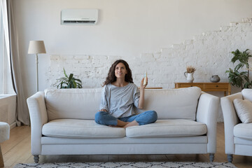 Attractive Hispanic woman holds remote controller turns on AC set comfort temperature in modern...