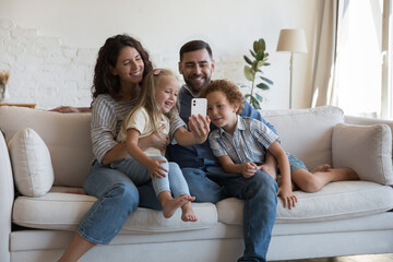Happy parents with little 5s children spend leisure using smartphone. Couple and siblings have fun...