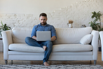 Handsome serious man sits on sofa with computer on laps, spend time at home web surfing information, use modern tech, make order on internet. Freelancing, telework, new software learning, tech concept