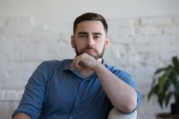 Pensive millennial man sit on couch in living room staring at camera. Profile picture of handsome...