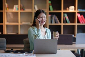 Beautiful Asian business woman chatting on the phone with customer and working on her laptop and taking note enthusiastically and happily in the office.