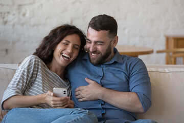 Carefree laughing 30s couple use cellphone relaxing on couch watch videos on internet feel happy spend weekend at home. Modern technology for fun, blogging, funniest websites for humour on web concept