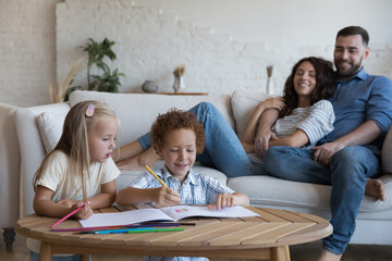 Full family spend free time on weekend together at own modern home, preschoolers children drawing in sketchbook with colored pencils, parents resting seated on cozy couch enjoy pastime and leisure