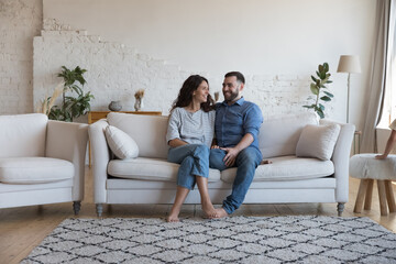 Smiling millennial loving couple sit on sofa enjoy pleasant talk, relaxing on weekend together at modern home, spend leisure or romantic date in cozy rented apartment. Tenancy, relationships concept