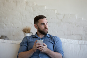 Pensive millennial bearded single man consider purchase sit on sofa hold smartphone distracted from gadget usage staring aside. Modern tech, remote communication, leisure using technologies concept