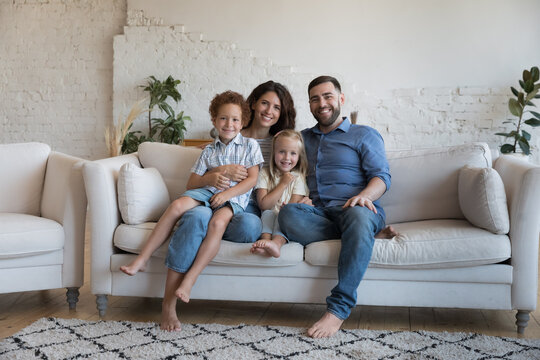 Happy well-being homeowners family portrait, unity concept. Young couple hugging preschoolers children smile relax on modern sofa in living room, enjoy weekend at new rented or own home