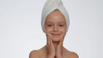 Lovely young child girl after bath in towel on head applying cleansing moisturizing cream. Teenager...