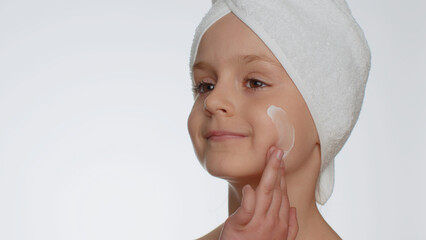 Young child girl after bath in towel on head applying cleansing moisturizing cream on white...