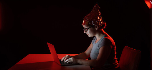 Paranoid caucasian woman typing on a computer wearing a tinfoil cap. Conspiracy theory.