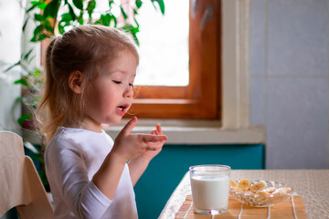 a little cheerful girl having breakfast with pieces of bananas and a glass of milk on the kitchen table in the morning. a child drinking a milk from a glass