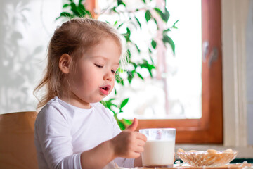 a little cheerful girl having breakfast with pieces of bananas and a glass of milk on the kitchen...