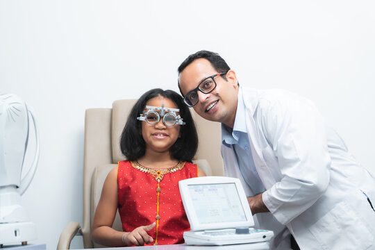 Happy Indian child girl doing examine eyesight with trial frame and lens with ophthalmologist or optometrist for eyes test at hospital or optometry clinic