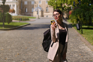 Fototapeta premium A cute young girl in glasses looks at the screen of a smartphone on a sunny day on the street of the old city.